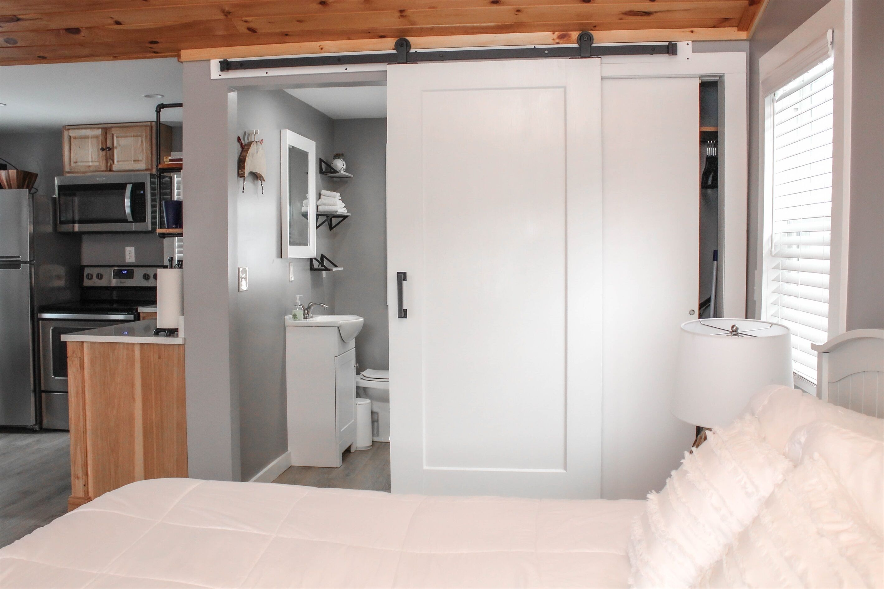 A bedroom with a bed, a nightstand, a white lamp, a sliding door to a bathroom, and a kitchenette with a microwave and stove.