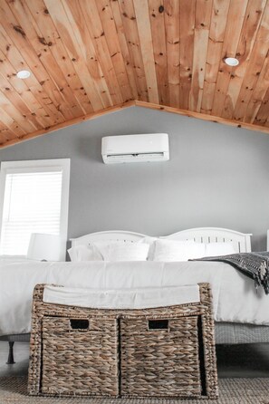 A bedroom with a sloped ceiling, a bed, a white air conditioning unit, and a wicker storage box. - Tranquil Cat Fish Cottage Getaway Point Breeze, NY (Kent)