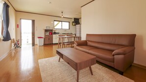 A modern living room with a brown leather sofa, a wooden coffee table, and a large window providing natural light. - Maru House (Uruma)