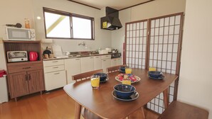 A dining area with a wooden table set for four, a kitchen with a microwave and toaster, and a sliding shoji screen. - Maru House (Uruma)