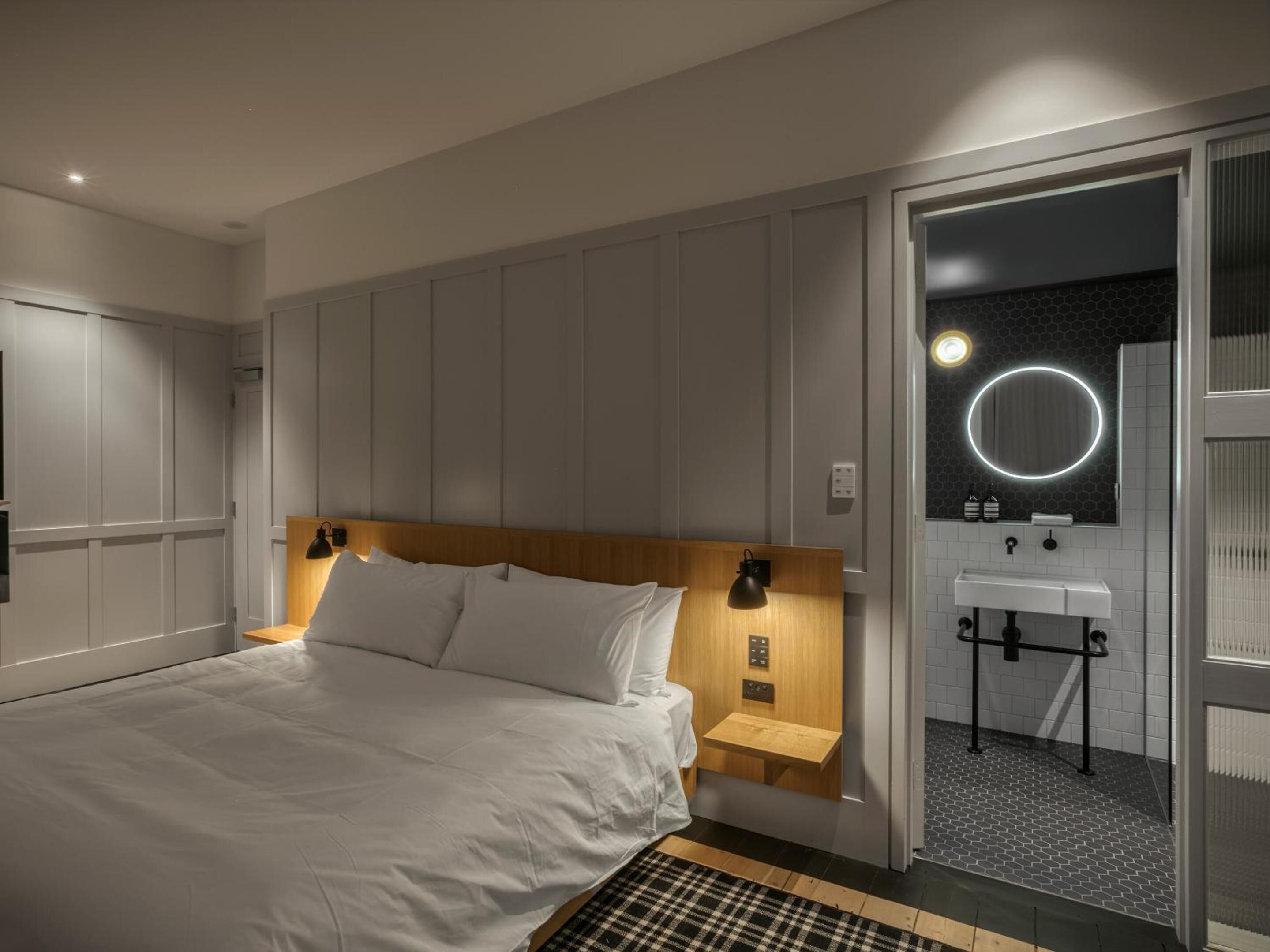 Une chambre d’hôtel moderne avec un grand lit, une tête de lit en bois, des lampes de chevet, une petite table de chevet en bois, un miroir rond avec un éclairage intégré et une salle de bain avec un lavabo et des toilettes visibles à travers une porte ouverte.