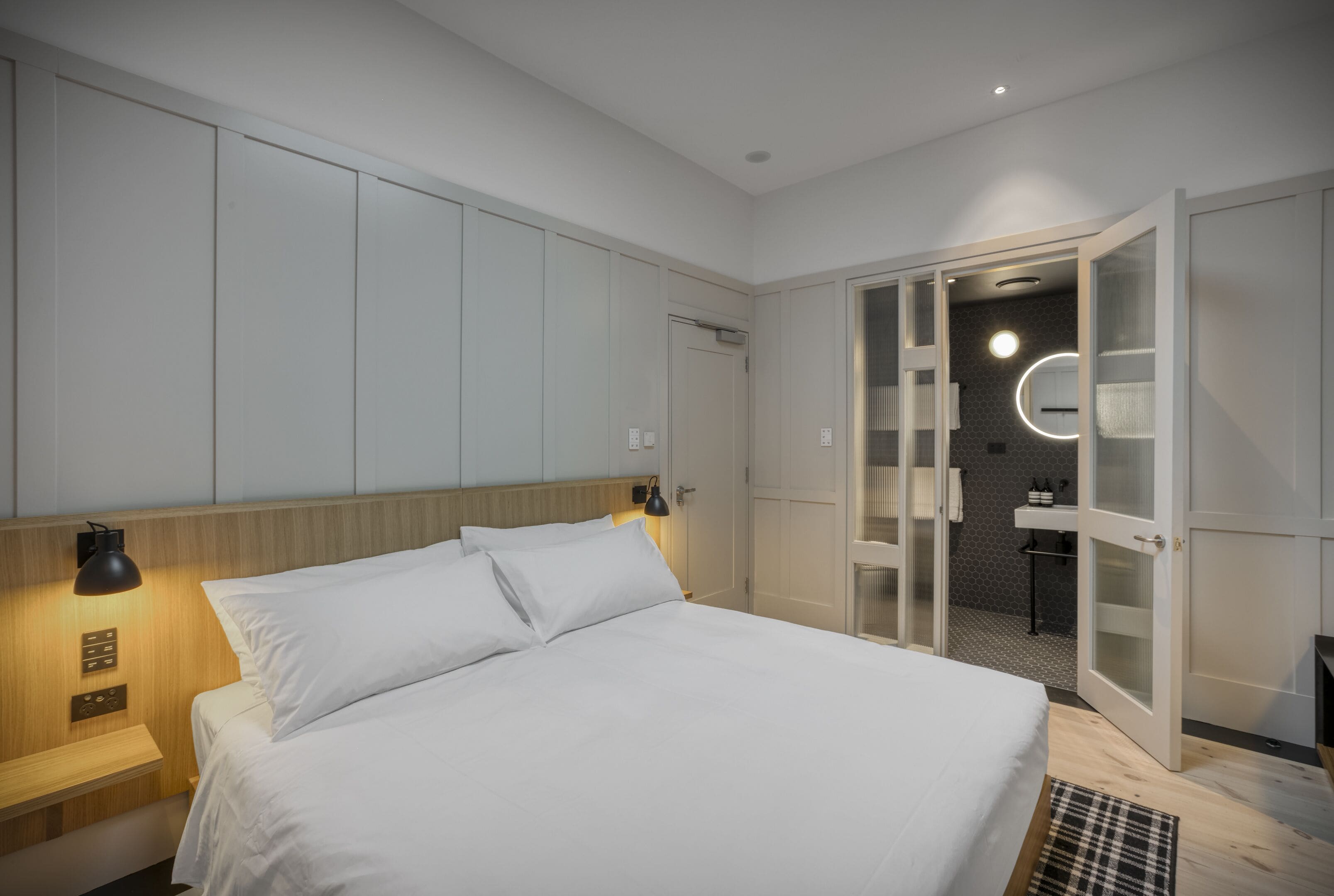 Una habitación de hotel moderna con una cama grande, cabecera de madera, mesita de noche con lámpara, y un baño visible a través de una puerta abierta.