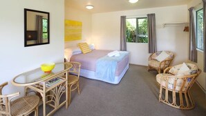 Een slaapkamer met een bed, twee rieten stoelen, een tafeltje met een schaal en een raam met gordijnen.