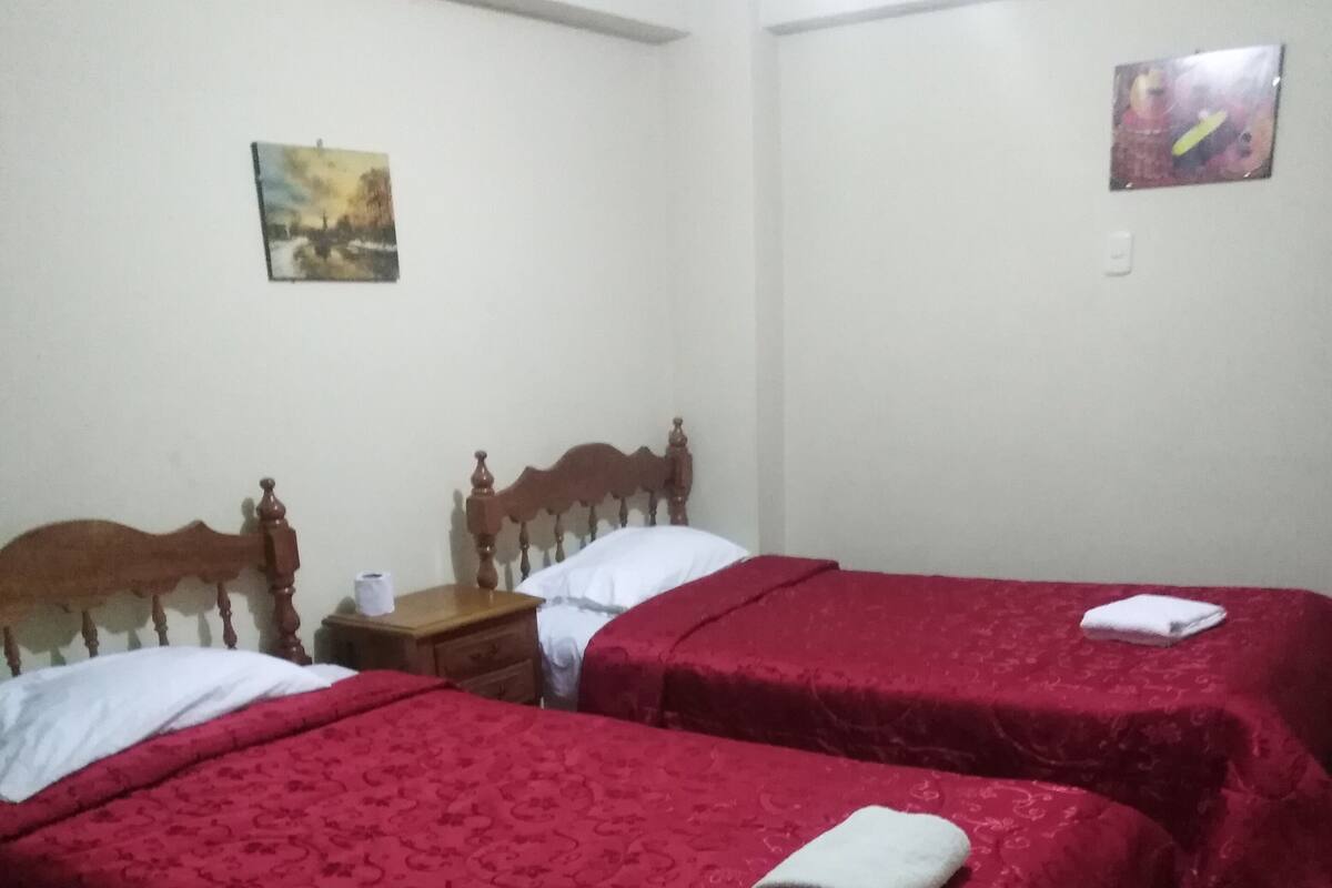 Een kamer met twee houten bedden, elk met een rode sprei en witte kussens. Er is een klein houten nachtkastje en een foto aan de muur.
