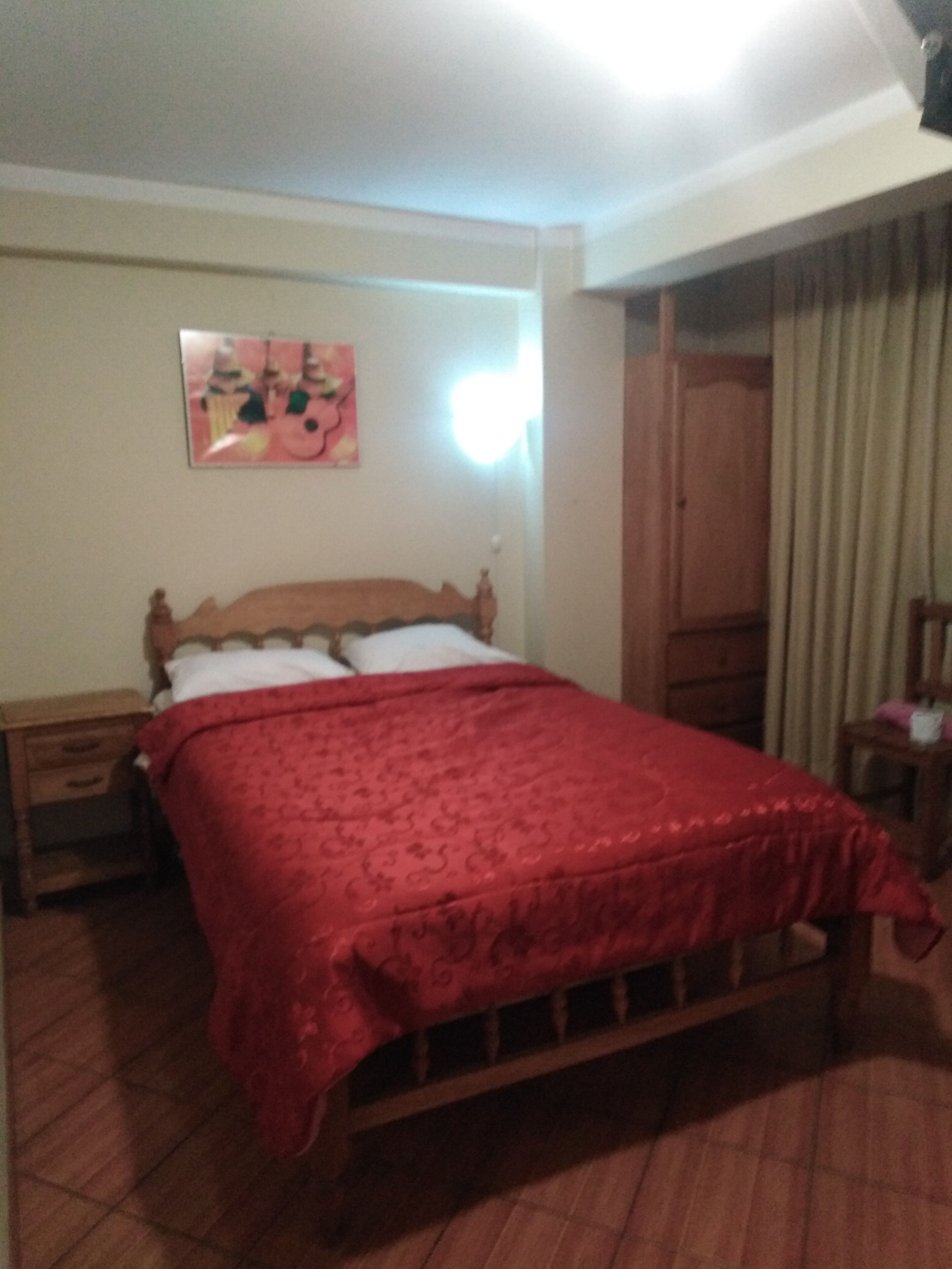 Un dormitorio con una cama de madera, una mesita de noche, una colcha roja, un cuadro en la pared y una ventana con cortinas.