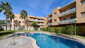 A swimming pool surrounded by a brick building with balconies, palm trees, and a clear sky.