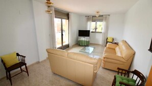 A living room with a beige sofa, a glass coffee table, and a TV on a stand. There are two chairs, one with a yellow cushion and another with a green cushion.