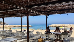 An outdoor seating area with tables and chairs, overlooking a beach with a rocky shoreline and the sea.