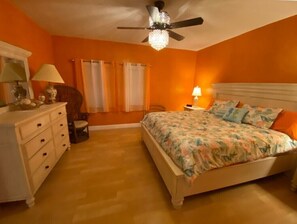 Une chambre avec un mur orange, un lit avec une literie à motifs floraux, une commode, une chaise, un ventilateur de plafond et une table de chevet avec une lampe.