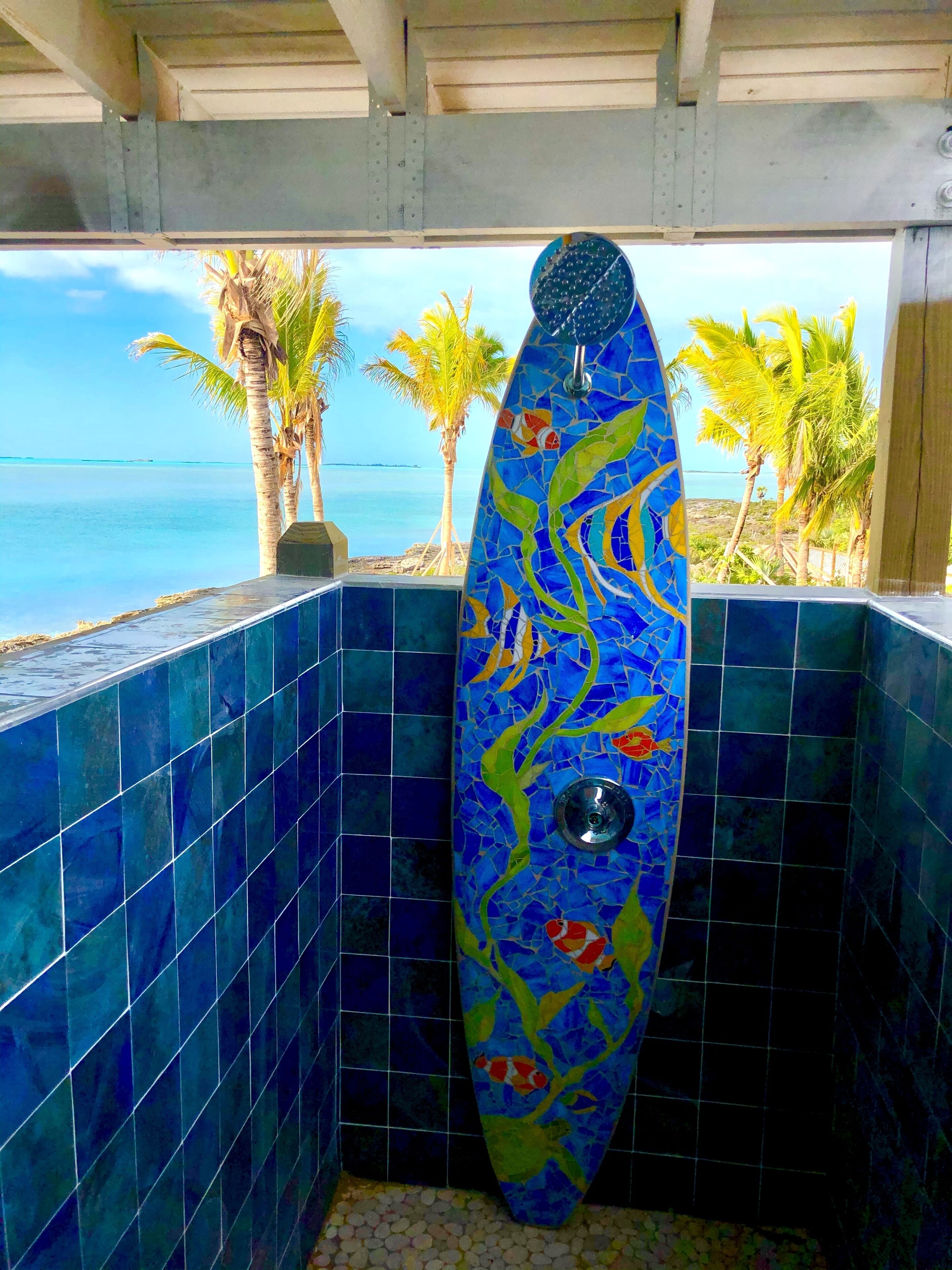 Une salle de bain avec des carreaux de mosaïque bleus, une cabine de douche et une vue sur l’océan et les palmiers.