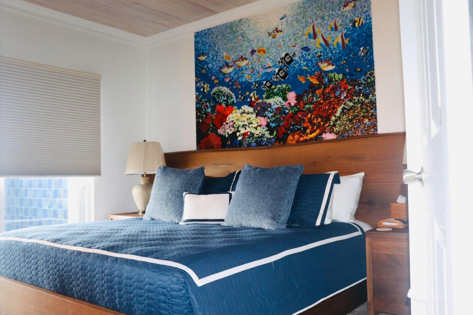 A bedroom with a large bed, a nightstand, a lamp, and a colorful ocean-themed painting on the wall.