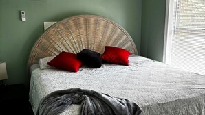 Una cama con cabecera de mimbre, manta gris y almohadas rojas y negras.