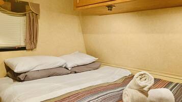 A small, cozy room with a bed, pillows, a striped blanket, and a rolled towel on the floor.