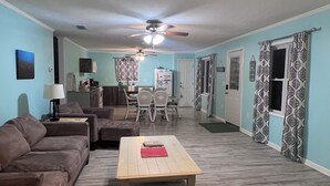 A spacious living room with a ceiling fan, a dining area with chairs, and a kitchen in the background.