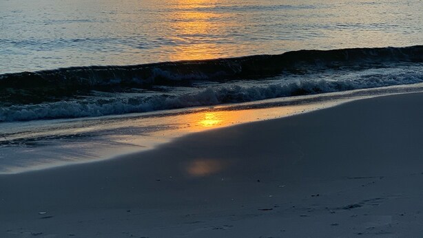 A beach at sunset with calm waves and a clear sky.