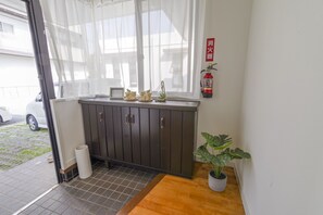 A room with a dark wooden cabinet, a small table with a potted plant, a fire extinguisher on the wall, and a window with sheer curtains. - Pine House in Araha (Chatan)