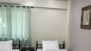Una habitación con dos camas individuales, cada una con ropa de cama blanca y una manta verde. Hay una mesita de noche negra entre las camas, y una ventana con cortina ligera.