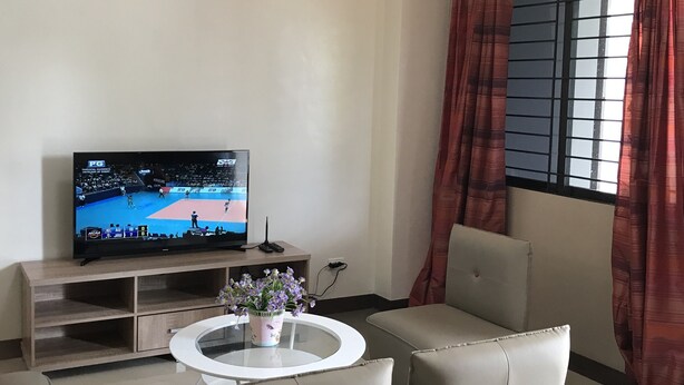 Una sala de estar con una televisión de pantalla plana, una mesa de centro de vidrio, dos sillones cómodos, una mesita con un jarrón de flores y una ventana con cortinas rojas.