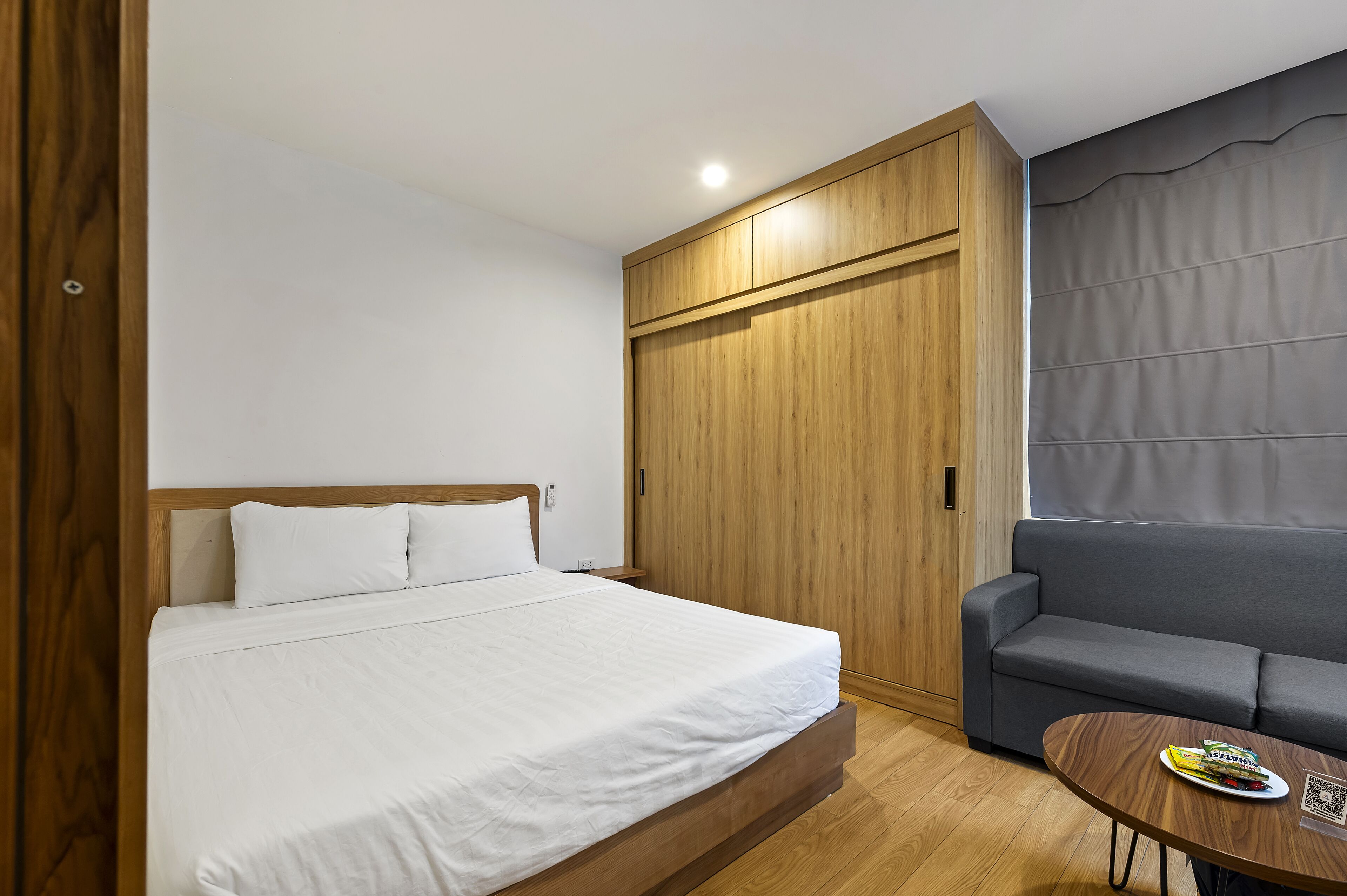 A bedroom with a bed, a wooden wardrobe, a grey sofa, and a small table with a magazine.