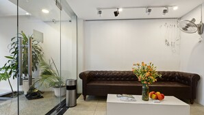 Ein Empfangsbereich mit einem braunen Ledersofa, einem weißen Couchtisch mit Blumen in einer Vase und einer Glastrennwand mit einem Topf und Pflanze.