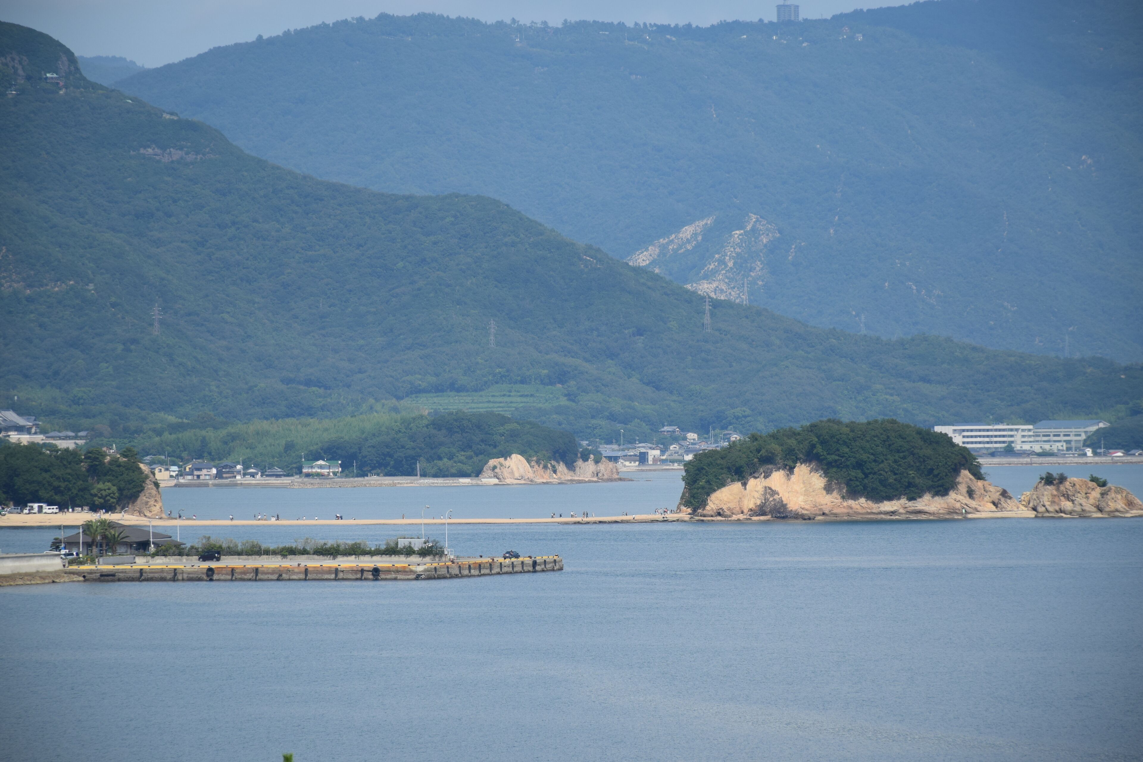 A coastal landscape with a pier extending into calm waters, surrounded by hills and a distant building.