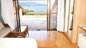 Une chambre avec un lit, un fauteuil en osier, une petite table et une vue sur une étendue d’eau, visible à travers une porte-fenêtre coulissante.