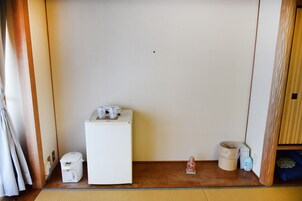 Una habitación pequeña con un refrigerador blanco, un basurero pequeño, un dispensador de agua y un cojín azul sobre un estante de madera.