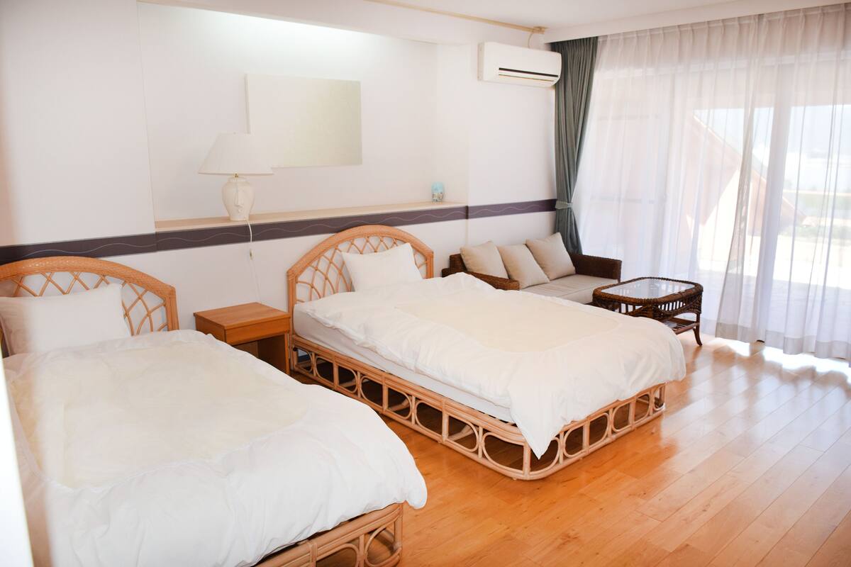 Ein Hotelzimmer mit zwei Holzbett, einem kleinen Tisch, einer Couch und einem großen Fenster mit durchsichtigen Vorhängen.