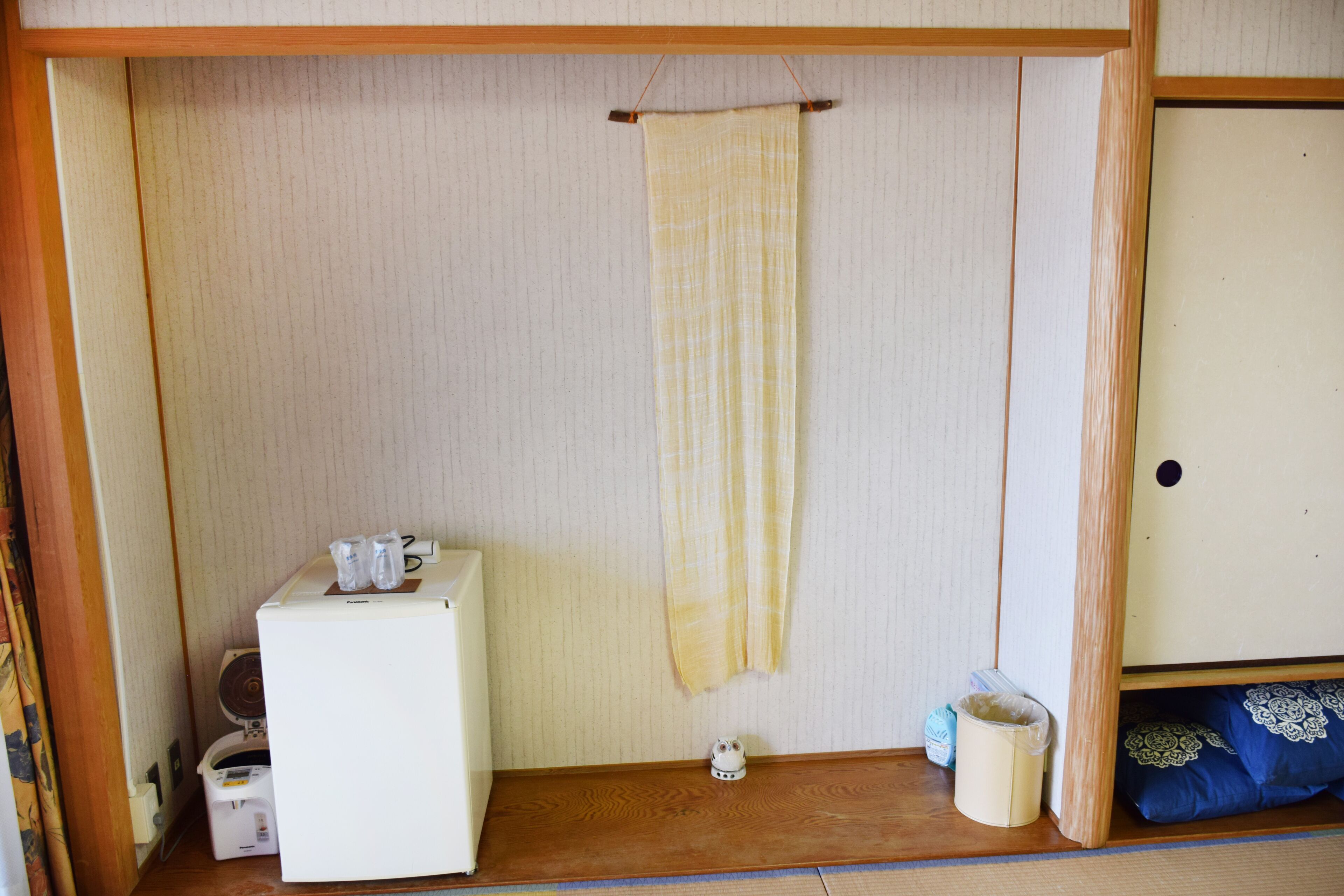 Een kleine, compacte kamer met een houten vloer, een witte koelkast, een kleine verwarming, een hangend gordijn en een blauw kussen op een laag bed.