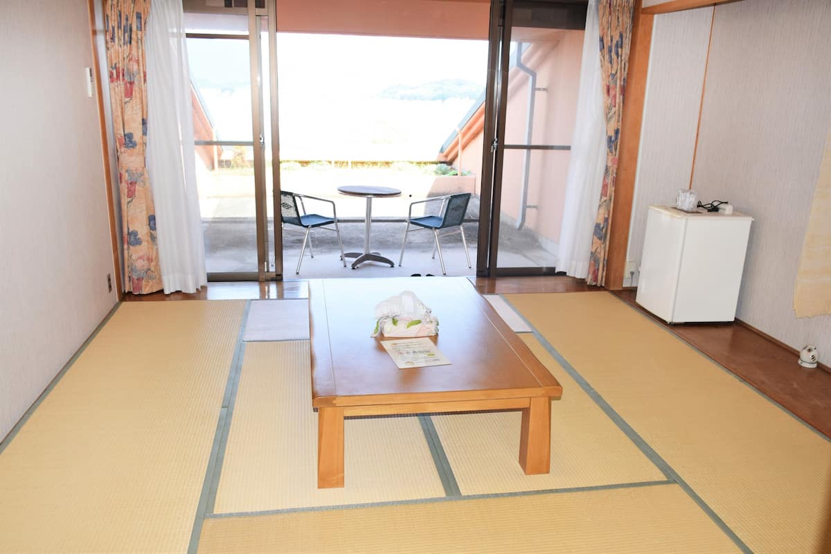 Ein Zimmer mit Tatami-Boden, einem Holztisch mit einer Tasche und einer Speisekarte, zwei Stühlen und einer Schiebeglasdoor, die zu einem Außenbereich mit Tisch und Stühlen führt.