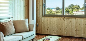 A room with a wooden paneled wall, a window with a view of houses and trees, a light-colored sofa, and a small table with items on it. - J&J Pension (Seogwipo)