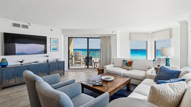 A modern living room with a large TV, comfortable seating, a coffee table, and a view of the ocean.