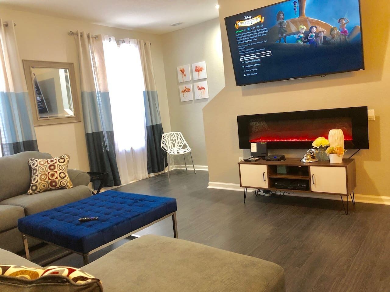En stue med et fladskærms TV ophængt på væggen, en sofa, en blå skammel og et lille skab med pynteting.