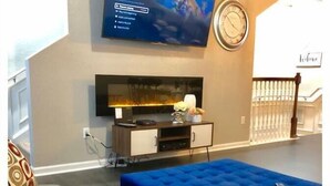 A living room with a flat-screen TV mounted on the wall, a clock, and a blue ottoman. - Tidecrest 144 · 3 Suites Near Convention, Universal, Disney (Orlando)