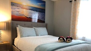 A neatly made bed with a headboard, a lamp on a bedside table, and a painting of a beach scene on the wall. - 6149 Sandcrest · Next TO Universal. 8 Beds. Pool. Very Clean (Orlando)