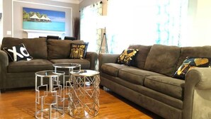 A living room with a brown sofa, two small round tables, and a painting of a tropical scene on the wall. - 6149 Sandcrest · Next TO Universal. 8 Beds. Pool. Very Clean (Orlando)