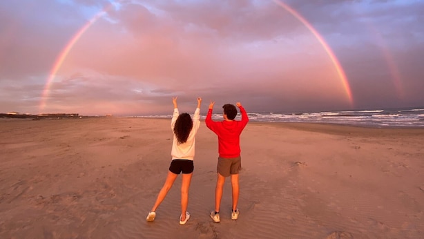 Deux personnes se tiennent sur une plage, et un arc-en-ciel apparaît dans le ciel.