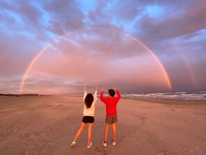 Two people standing on a beach with a rainbow in the sky.