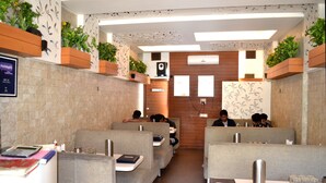 A restaurant with booths, tables, and a counter. There are plants on the walls, a sign for a wash room, and a speaker on the wall. - Hotel Swagat (Abu Road)