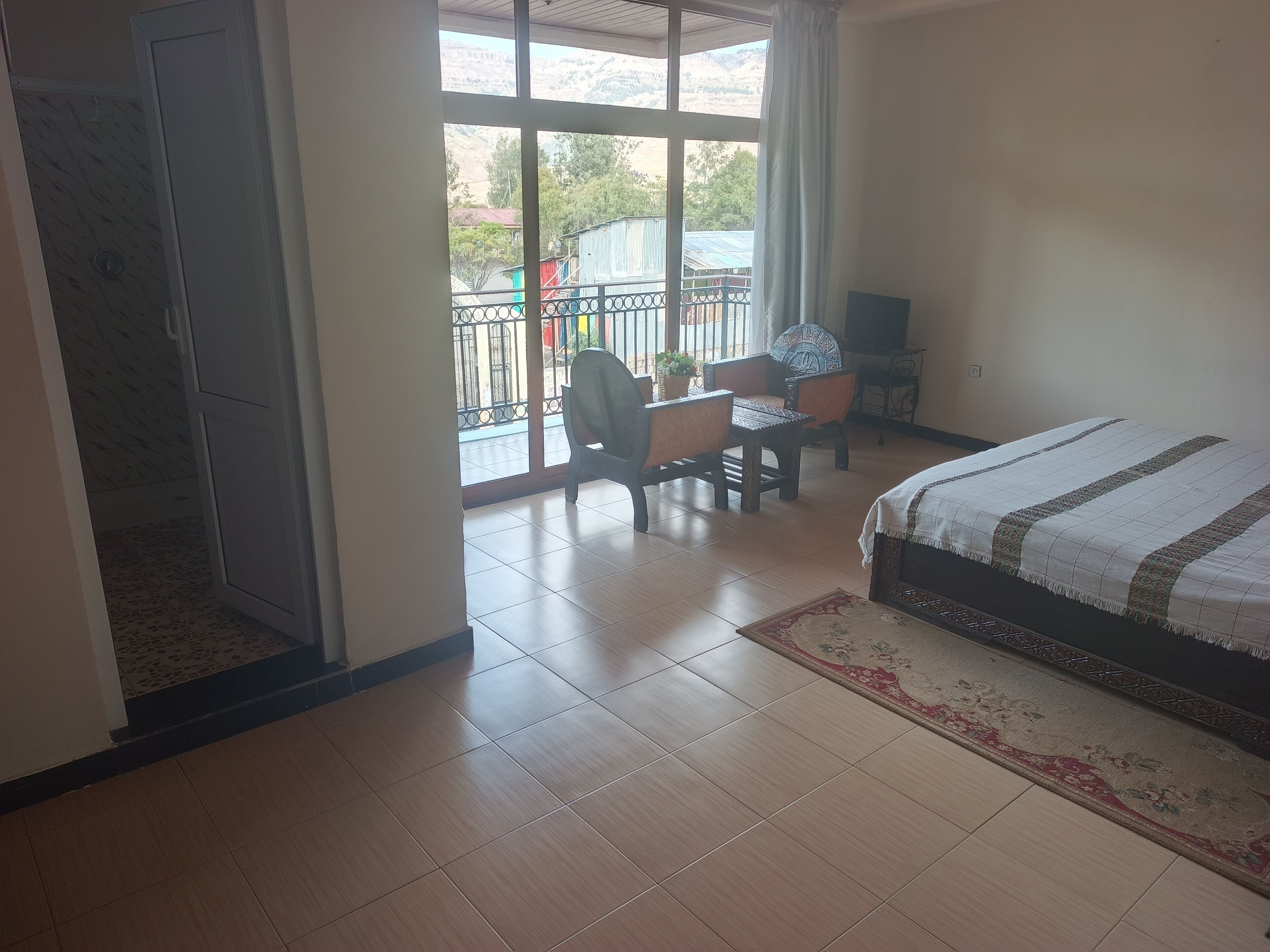 A bedroom with a bed, a desk, and a chair. There is a balcony with a view of the outside.