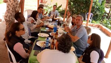 Een groep mensen zit aan tafel met eten en drinken, onder een overkapping met uitzicht op groen.