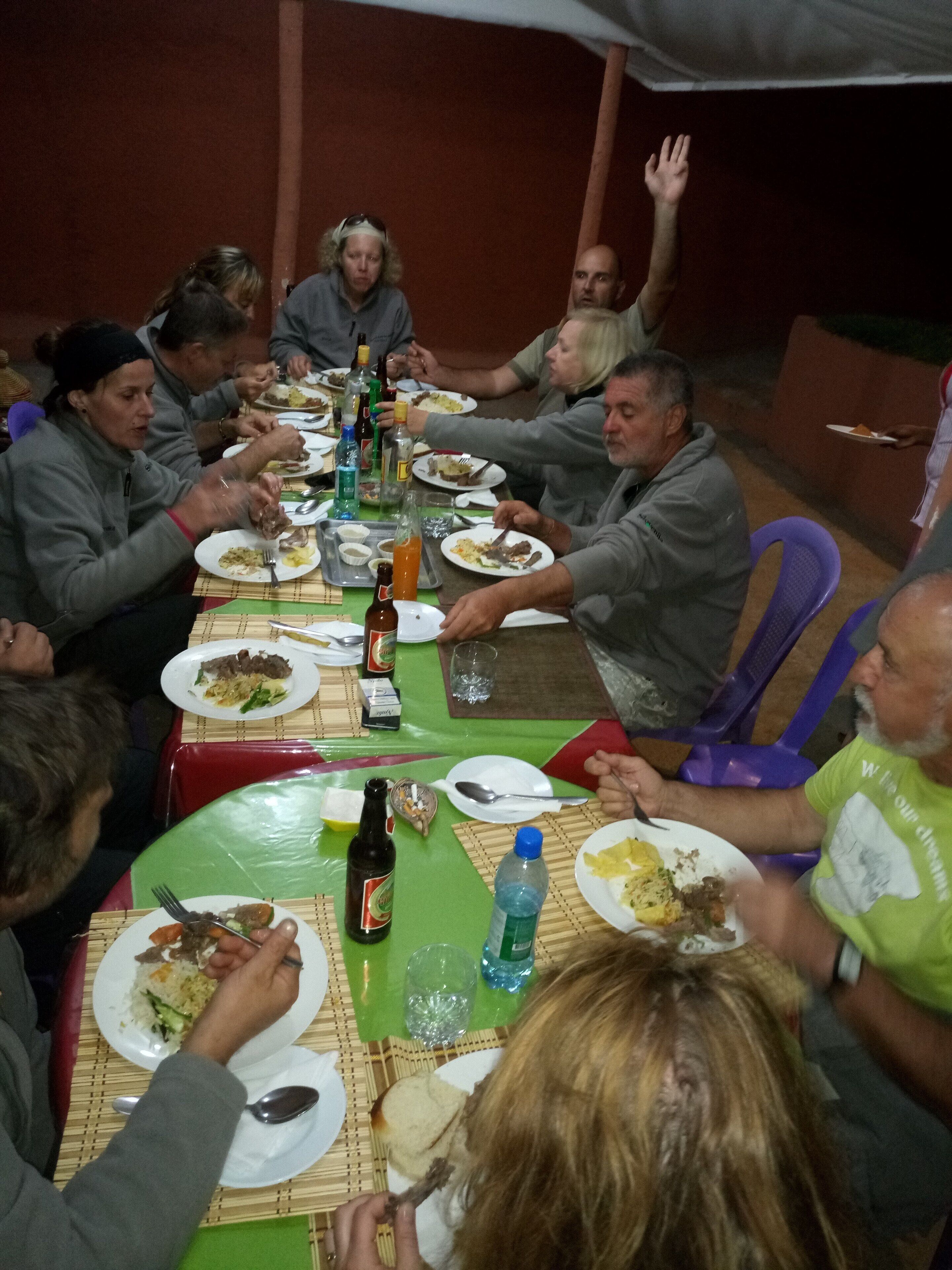 A group of people seated around a table with plates of food, bottles, and a bottle of beer.