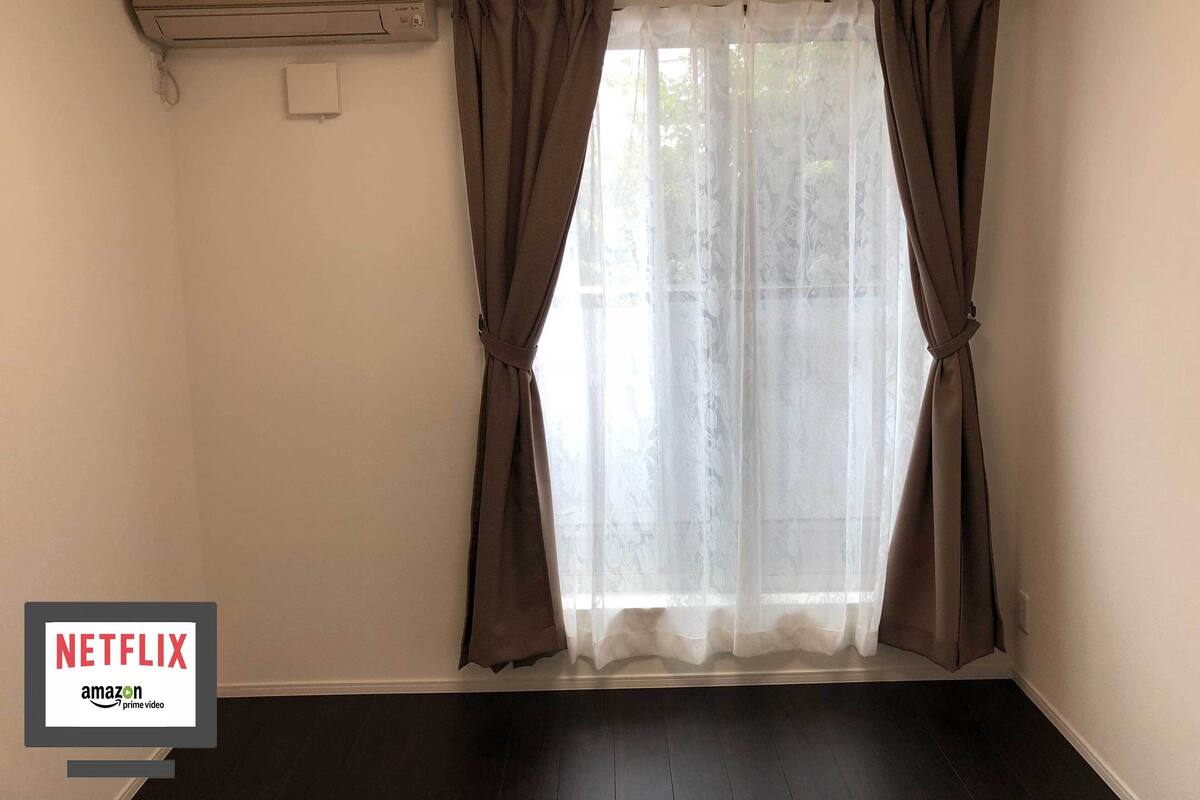 A room with a window, brown curtains, a white air conditioning unit, and a dark wooden floor.