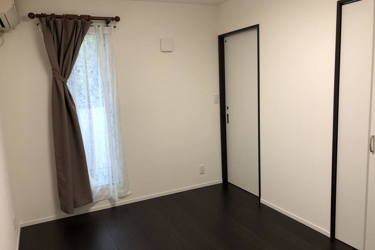 A room with dark wooden flooring, a closed door, and a window with sheer curtains.