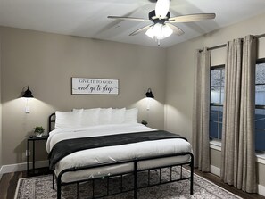 Un dormitorio con cama, ventilador de techo, dos lámparas de noche, ventana con cortinas y una cita motivadora en la pared.