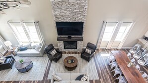 A spacious living room with a stone fireplace, a large flat-screen TV, a sofa, two armchairs, a coffee table, and a dining area with a long wooden table and white chairs.