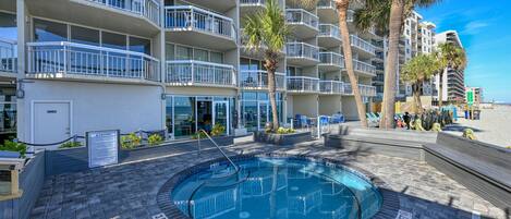 A hotel with a circular pool surrounded by a brick patio, palm trees, and a beach view.