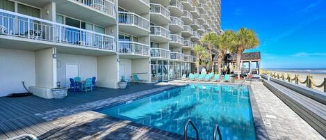 A pool area with a clear view of the ocean, a building with balconies, and lounge chairs.
