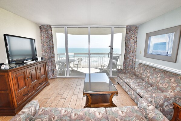 Uma sala de estar com sofá, mesa de centro, televisão e varanda com vista para o mar.