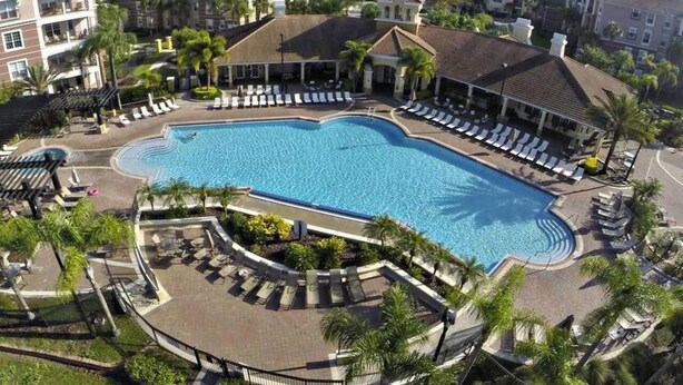 A large swimming pool surrounded by lounge chairs and palm trees, with multi-story buildings in the background. - Tideview 20 · Walk TO Convention! Near Universal & Disney (Orlando)